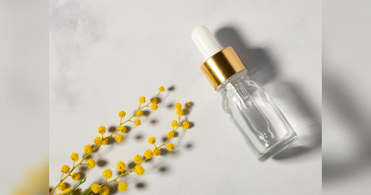 Frasco transparente com conta-gotas ao lado de flores amarelas em fundo claro.