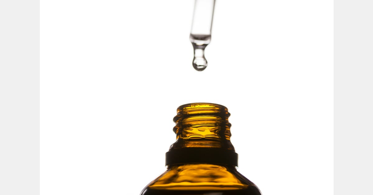 Conta-gotas com gota sobre frasco âmbar aberto em fundo claro.